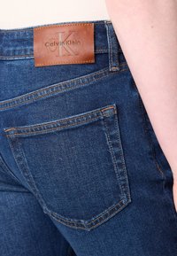 Jeans de ganga azul-escuro com costura laranja, apresentando um rótulo de pele castanha da Calvin Klein na cintura e dois bolsos traseiros.