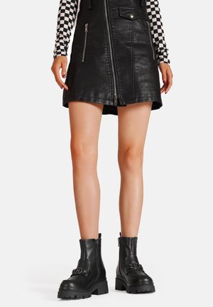 Stivali alla caviglia in pelle nera con suole spesse, hardware decorativo, cerniere laterali e superficie liscia. Indossati con un top a scacchi bianco e nero.