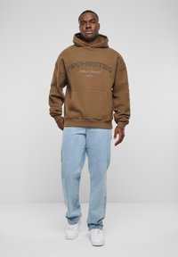 Brauner Hoodie mit dem Text "VERBOTEN" und einer Kängurutasche, kombiniert mit hellblauen Jeans und weißen Sneakers. Einfaches Design mit gerippten Bündchen.