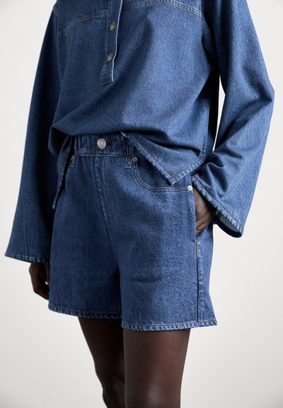 Denim šorti vidēji zilā krāsā ar brīvu piegriezumu, augstu jostasvietu un piecām kabatām. Satur kontrastējošu izšuvumu un vieglu nodilumu.