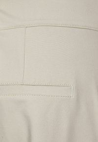 Läheltä otettu kuva vaatteesta, jossa on vaalea beige kangas, sileä pinta, näkyvät ompeleet ja yksi uhkeasti viimeistelty tasku.