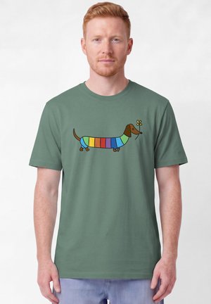 Homme portant un t-shirt vert avec un teckel rayé coloré tenant une fleur jaune dans sa bouche, debout sur fond blanc.