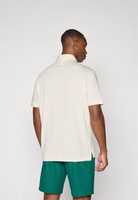 Cămașă polo crem cu mâneci scurte, guler ribbed, fante laterale și o textură netedă, combinată cu pantaloni scurți verzi. Vedere din spate.