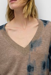 Pull col en V dans un ton beige et bleu marine, présentant une texture tricotée et des motifs abstraits teints sur le tissu.