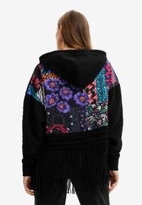 Sudadera con capucha que presenta un diseño de patchwork con patrones florales, estampados de animales y acentos de flecos. Fabricada en tejido negro con colores vibrantes.