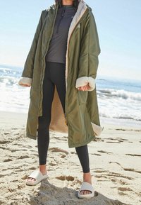 Oliven grøn lang parka med pelsforet manchetter og inderside, lynlåsåbning foran, kombineret med sorte leggings og beige slidesandaler på sand.