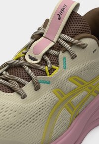 Close-up of beige and brown ASICS sneaker with pink tab, yellow logo, teal stitching, and brown-and-white speckled laces.