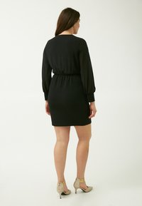 Schwarzes Kleid mit langen, gepufften Ärmeln, taillierter Taille und engem Rock. Wird mit beigen High Heels mit goldenen Akzenten getragen.