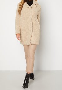 Beige, fluffy frakke med knaplukning, krave og sidelommer, båret over cremefarvede bukser og spidse sorte ankelstøvler.