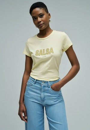 Mujer con camiseta amarilla clara "SALSA JEANS" y vaqueros azul claro, posando con una mano en el bolsillo contra un fondo liso.