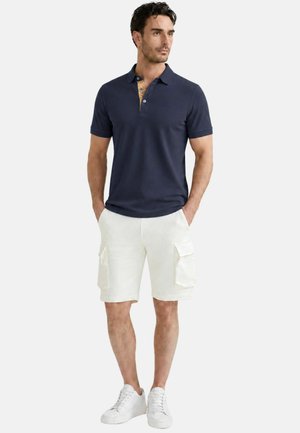 Uomo con polo blu navy, pantaloncini cargo bianchi e scarpe da ginnastica bianche, in piedi con le mani in tasca guardando di lato.