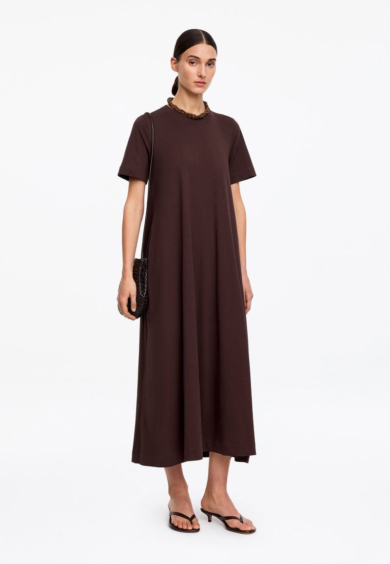 Femme portant une robe maxi ample à manches courtes marron foncé, portant un petit sac texturé et des sandales noires à talons.