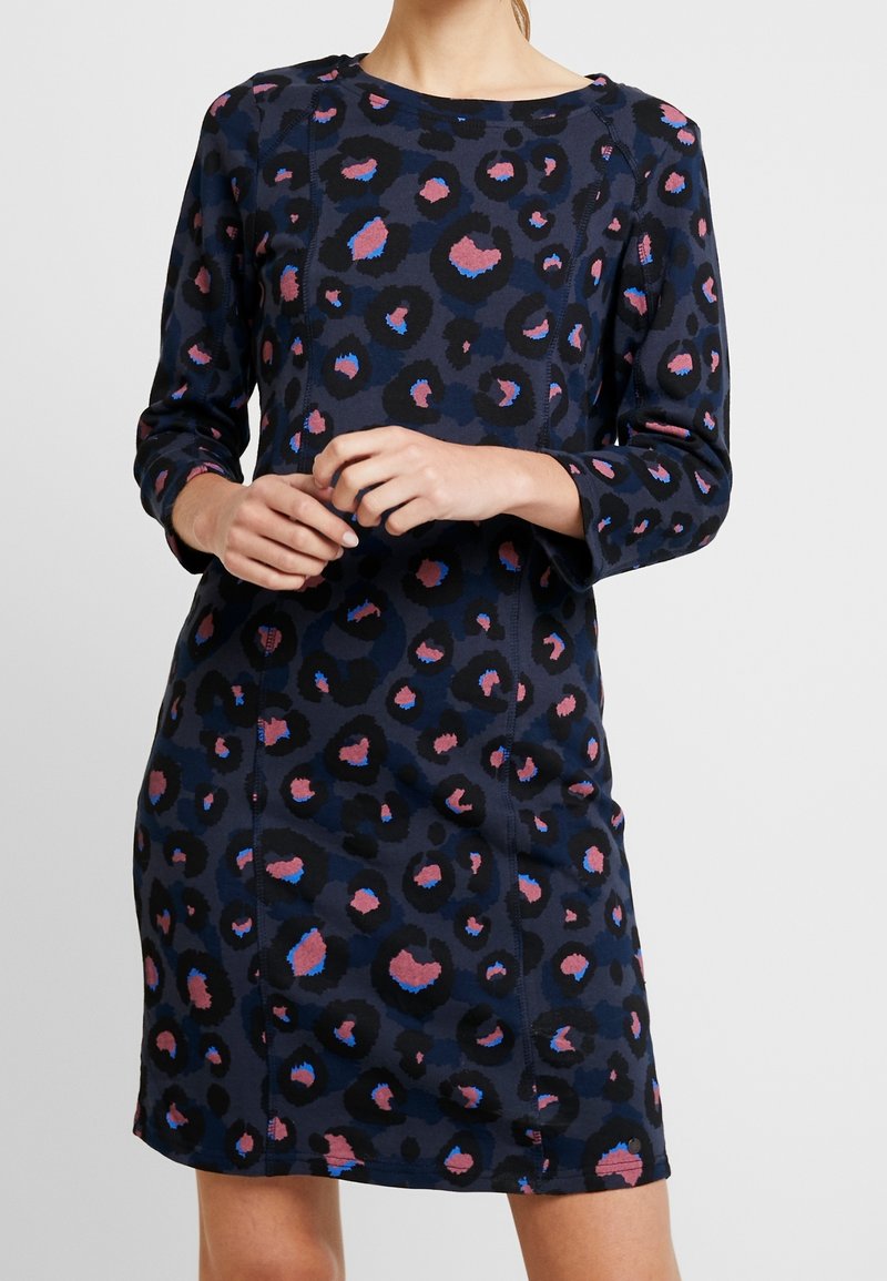 Femme portant une robe bleu marine à imprimé léopard rose et bleu, manches longues, encolure ronde, mains jointes à la taille.