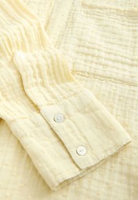 Light yellow textured fabric with a subtle dotted pattern. Features a button cuff with two pearl-like buttons, depicting a soft, breathable design.