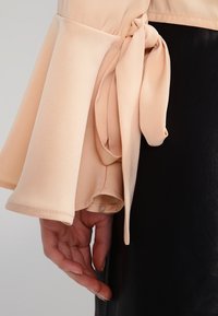 Manica di una blusa color pesca con design a volant e un nastro annodato al polso, abbinata a un indumento nero texturizzato.