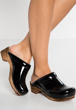 Gambe con zoccoli in pelle verniciata nera con suola in legno su una superficie grigio chiaro, un piede leggermente sollevato.