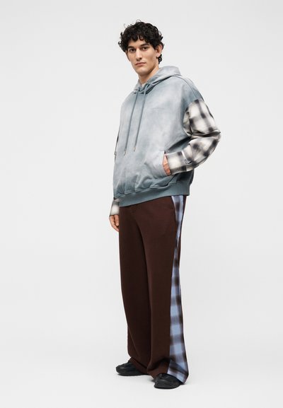Νέος άνδρας που φοράει γκρι φούτερ tie-dye με καρό μανίκια και καφέ παντελόνια με ριγέ καρό στο πλάι, στέκεται με τα χέρια εν μέρει στις τσέπες.