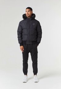 Mann iført svart pufferjakke med pelshett, svart cargo-joggebukser og hvite sneakers, stående mot en lys enkel bakgrunn.