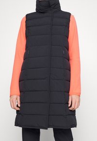 Svart pufferväst med hög krage, vertikal quiltdesign, två sidofickor och medellång längd, tillsammans med en korallfärgad långärmad topp.