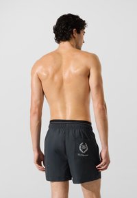 Homem sem camisa com cabelo cacheado, vestindo calções de banho pretos com um bolso nas costas e um logótipo de grinalda branca com texto sobre um fundo simples.