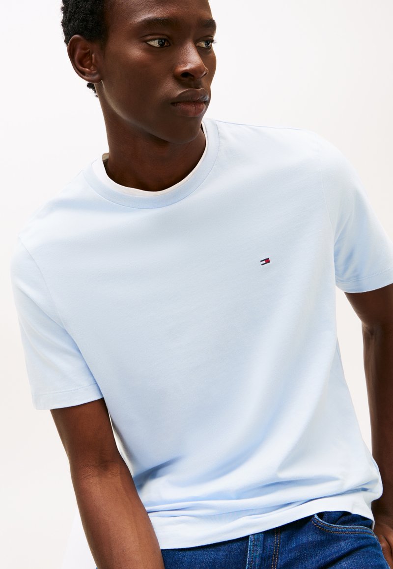 Fiatal férfi világoskék rövid ujjú pólót visel, melyen kis Tommy Hilfiger logó található a mellkason, kék farmerral párosítva.