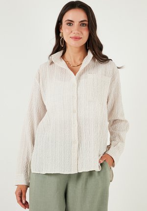 Light beige button-up shirt with a textured fabric, featuring long sleeves, a pocket on the left, and a collared neckline.