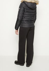 Chaqueta negra acolchada con capucha con borde de piel, diseño acolchado horizontal y un logotipo en la manga izquierda, combinada con pantalones anchos negros.