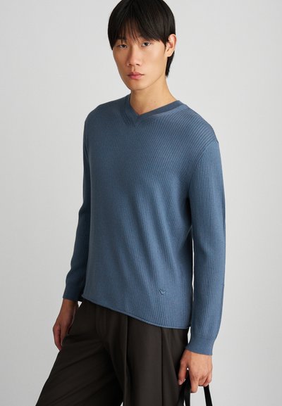 V-hals sweater i blødt, ribbet materiale; har en afdæmpet blå farve, lange ærmer og subtil branding ved kanten. Båret med mørke bukser.