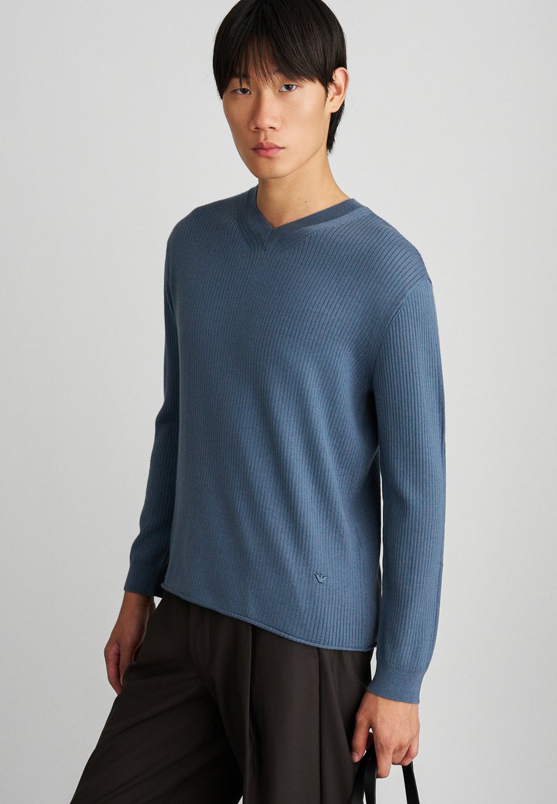 Πουλόβερ V με μαλακό, ραμμένο υλικό. Διαθέτει μια απαλή μπλε απόχρωση, μακριά μανίκια και διακριτική επωνυμία στο στρίφωμα. Συνδυάζεται με σκούρα παντελόνια.