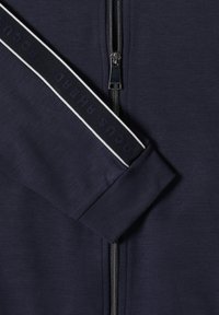 Granatowa kurtka na zamek błyskawiczny o gładkiej fakturze, z srebrnym zamkiem i czarnym paskiem z białym napisem na rękawie.