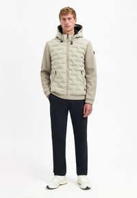 Man staat in een beige pufferjack met capuchon, zwarte broek en witte sneakers tegen een effen witte achtergrond.