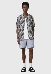 Cămașă cu imprimeu tropical în alb și negru cu mâneci scurte, purtată peste un top alb texturat, asortată cu pantaloni scurți gri deschis și sandale negre cu șosete albe.