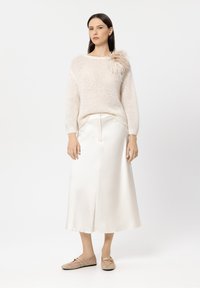 Hellbeiger Strickpullover mit strukturiertem Material und einem Federakzent an der linken Schulter, kombiniert mit einem cremefarbenen Satin-Midikleid und beigen Slippers.