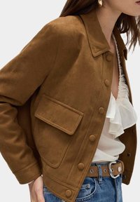 Giubbotto di suede marrone con colletto, bottoni a scatto e tasca a patta. Indossato con un top bianco arricciato e jeans blu. Texture liscia.