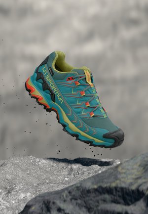Teal hiking shoe with vibrant orange and yellow accents, mesh upper, robust sole, and contrasting textured patterns for traction, suspended above rocky surface.