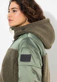 Grön hybridjacka med en fluffig fleeceutvändighet, glänsande nylondelar, foderhuva och en svart logotyp på ärmen.