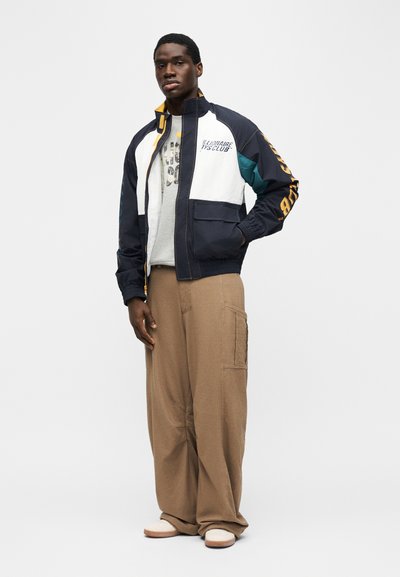 Άνδρας που φοράει λευκό και ναυτικό μπλε μπουφάν με φερμουάρ και το κείμενο «Billionaire Boys Club», μπεζ φαρδιά παντελόνια και λευκά αθλητικά παπούτσια, στέκεται με το ένα χέρι στην τσέπη.