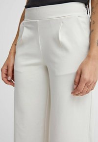 Pantaloni bianchi a gamba larga con una texture liscia, caratterizzati da pieghe frontali e una vita alta. Mano visibile che poggia delicatamente sul tessuto.