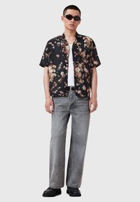 Bărbat îmbrăcat cu o cămașă scurtă cu imprimeu floral negru, un tricou alb pe dedesubt, blugi gri largi, pantofi negri, ochelari de soare și cercei din argint, stând pe un fundal simplu.