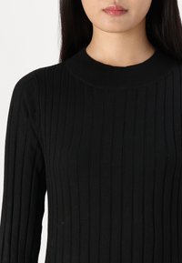 Sweater preto canelado com gola redonda, apresentando linhas verticais e uma textura suave. O tecido é liso e ajustado, acentuando a silhueta.