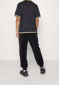 Uomo in piedi con le spalle alla fotocamera, indossa pantaloni neri con striscia laterale, scarpe da ginnastica nere e una maglietta nera a maniche corte con bordi a righe.
