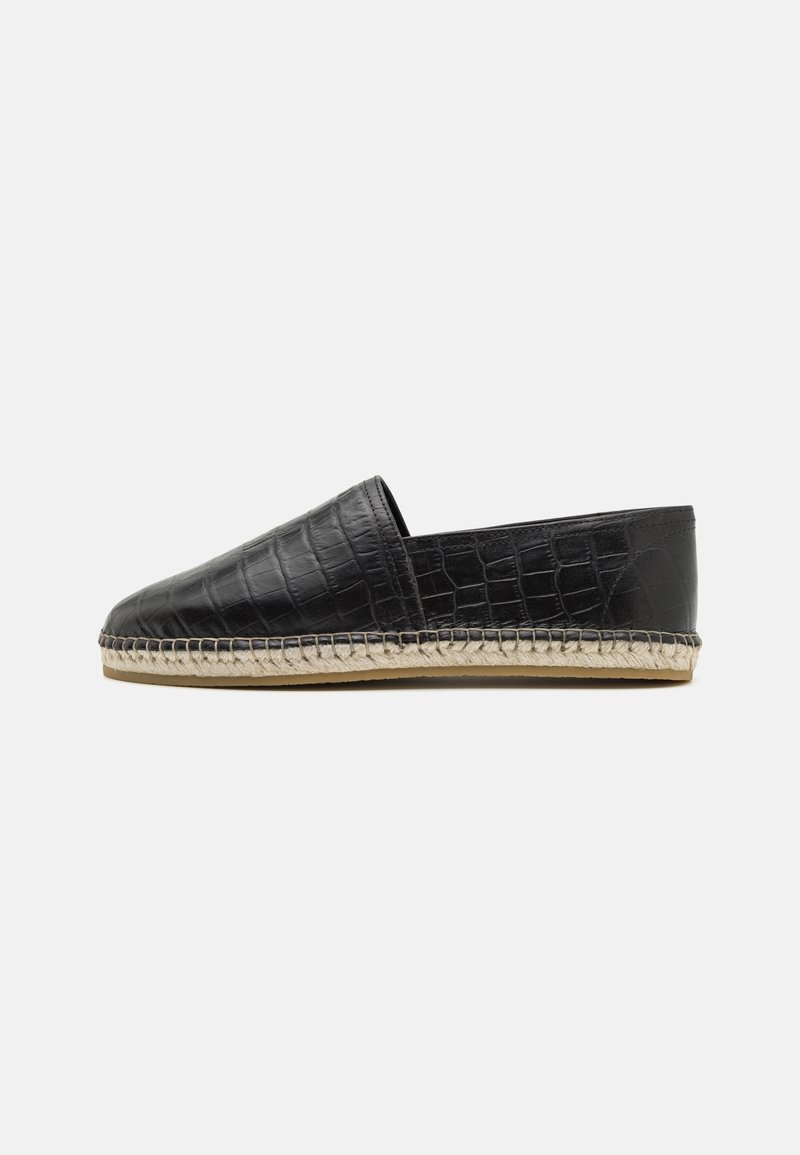 Chaussure noire à enfiler avec texture crocodile, semelle en jute tissée et semelle plate en caoutchouc, montrée de profil sur fond blanc.