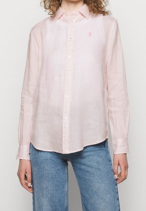 Žena oblečená v světle růžové košili s dlouhým rukávem na knoflíky s malým vyšitým logem, doplněná modrými džínami.