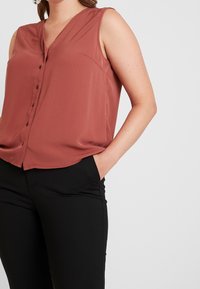 Blouse sans manches couleur rouille avec un col en V, présentant un devant boutonné et une seule poche poitrine. Assortie à un pantalon noir ajusté.