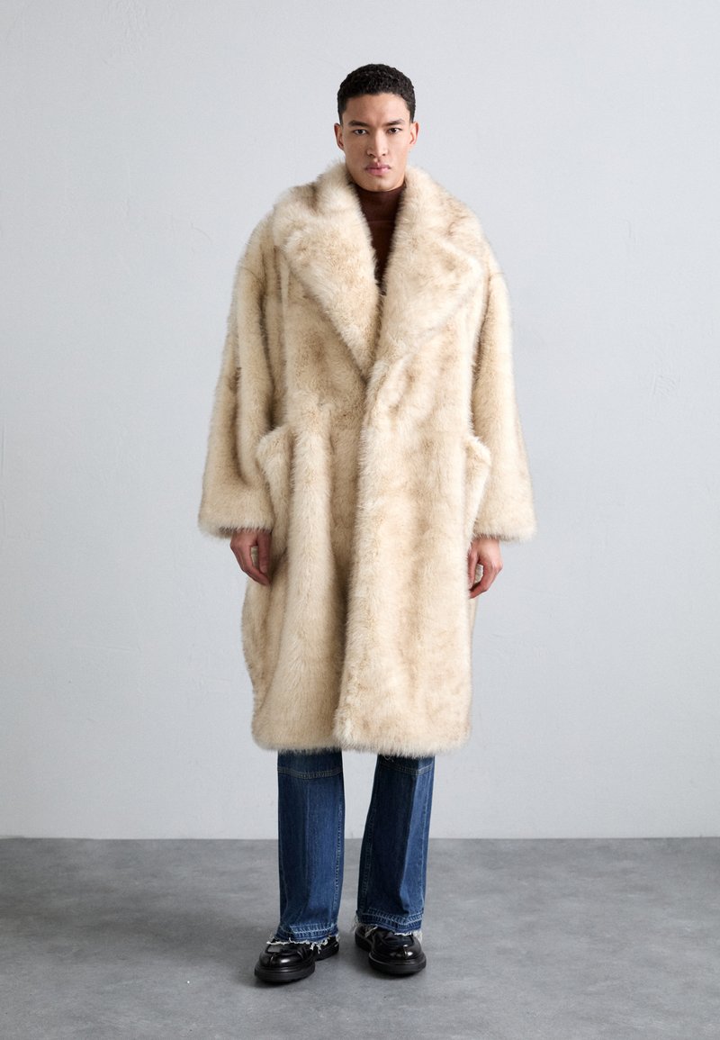 Šviesiai ruda dirbtinio kailio striukė su plačiais atlapais, per didelis dizainas ir gilūs kišenės, derinama su mėlynomis džinsomis ir juodais batais.