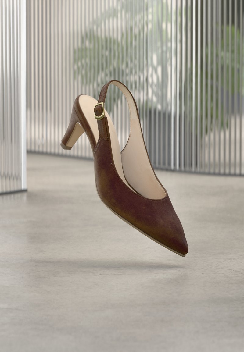 Καφέ γόβα με λουρί και ψηλό τακούνι από καστόρι, με μυτερή μύτη και χρυσή αγκράφα, αιωρείται πάνω από ένα γυαλισμένο τσιμεντένιο πάτωμα με θολά κάθετα πάνελ στο βάθος.