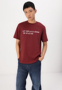 Maroon short-sleeve t-shirt with white text reading "LOT REPLAY B. DENIM RD 39 423 925," paired with blue corduroy pants. Smooth cotton fabric.