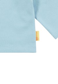T-shirt en coton bleu clair à manches courtes, avec un ourlet droit et une petite étiquette jaune sur le côté avec un logo circulaire.