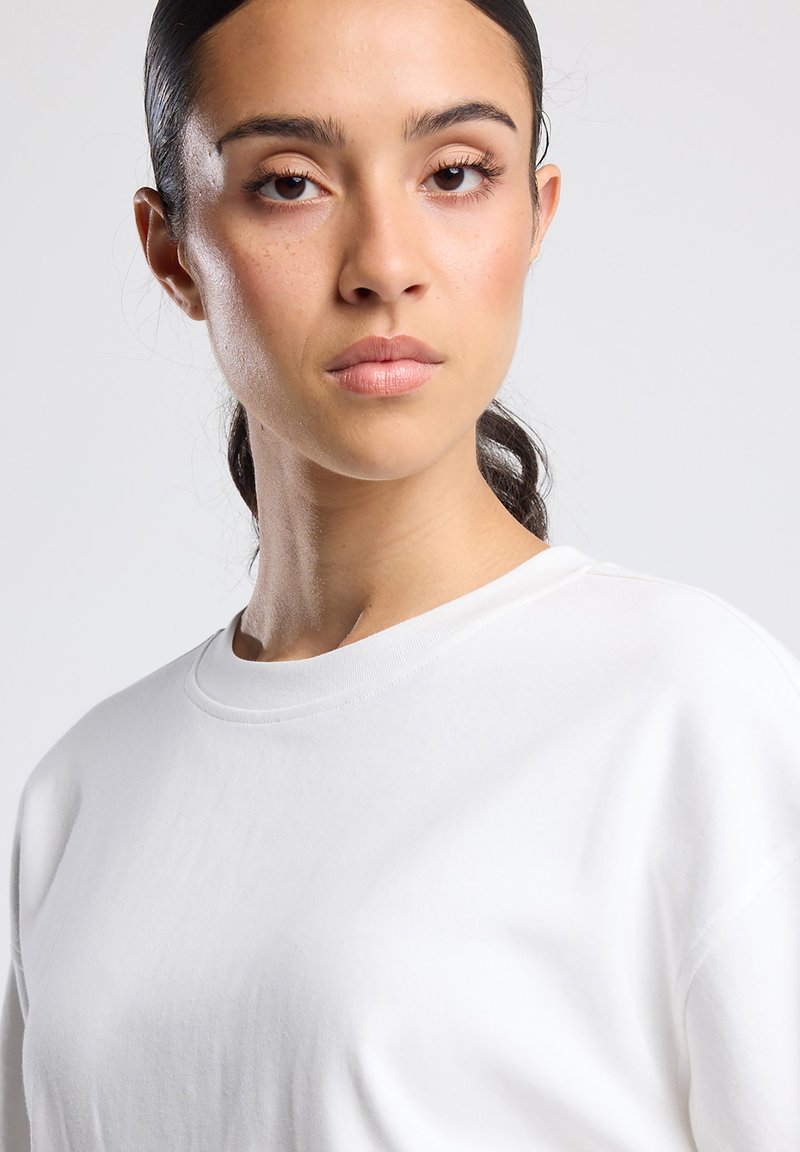 Young woman with dark hair tied back wearing a plain white crewneck shirt against a light gray background.