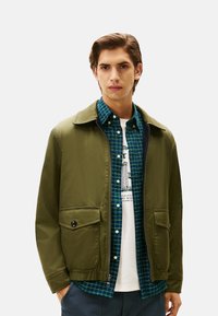 Olijfgroene lichte jas met kraag, rits sluiting en twee voorzakken. Draag het over een blauwgroene geruite blouse en een wit T-shirt met opdruk.